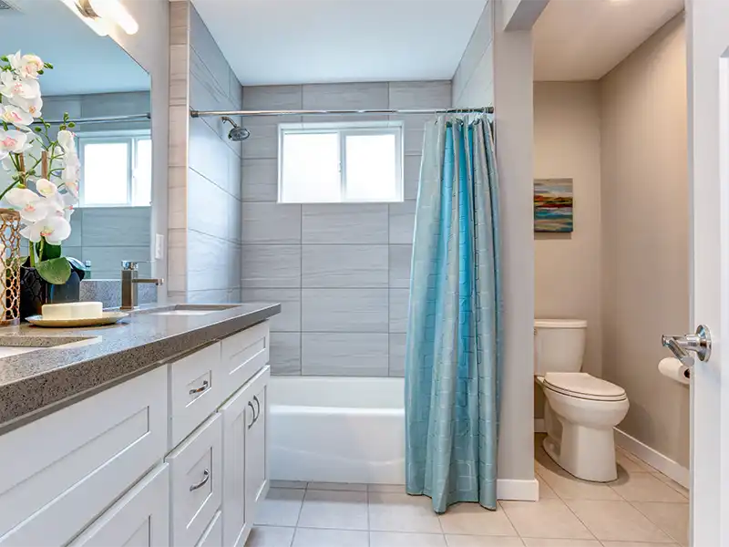 Modern bathroom with white vanity, gray tiled shower, blue curtain, and toilet in a neutral-toned room. sewage cleanup
