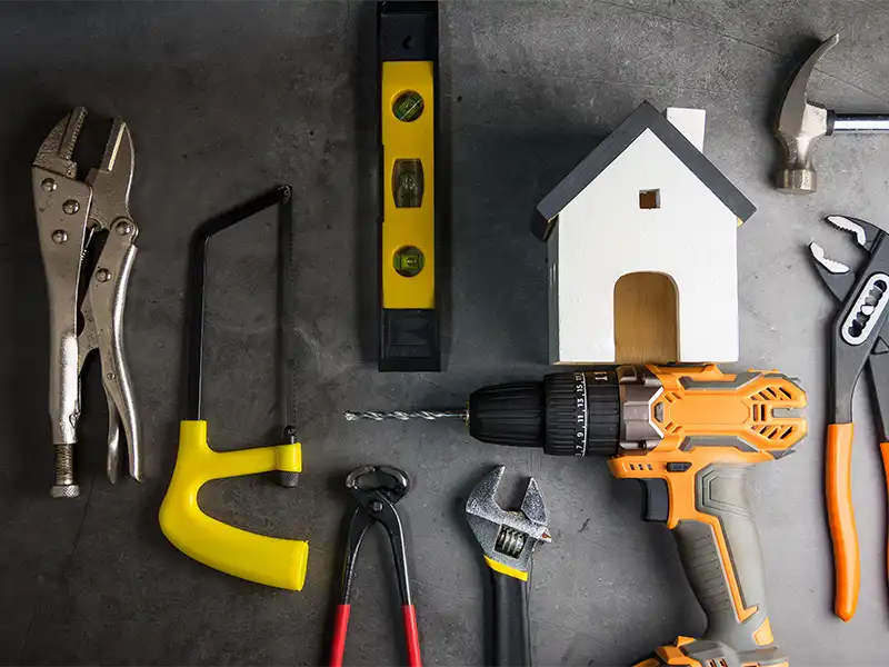 Various hand tools including a drill, hammer, wrench, and a small wooden house model on a gray surface.