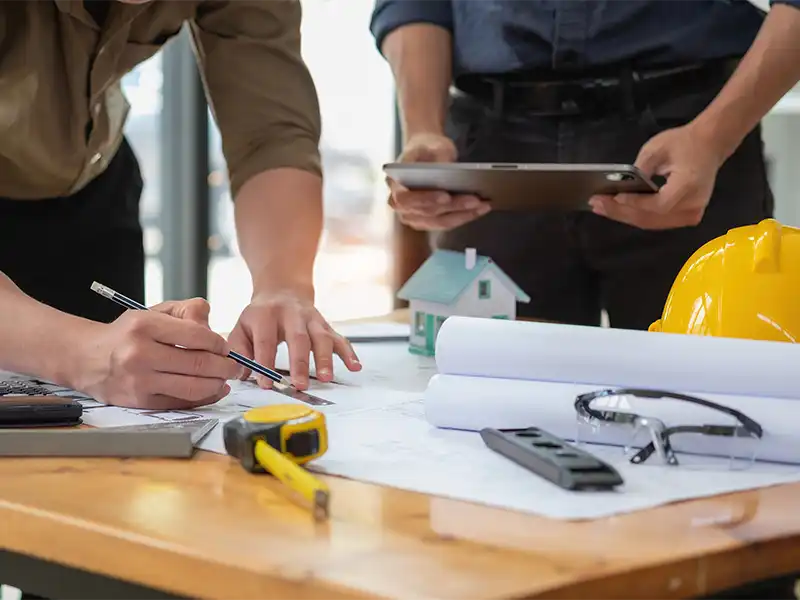 Two people reviewing architectural plans with tools, a model house, and a yellow hard hat on the table.