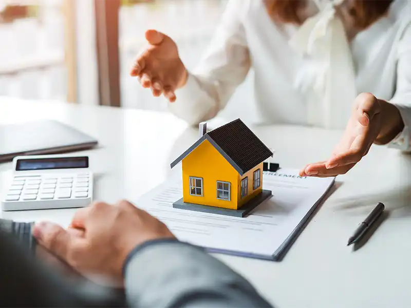 Two people discussing a small yellow house model on top of documents at a table.