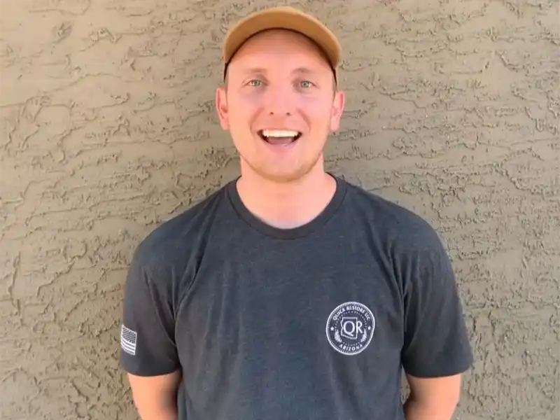 Man wearing a tan cap and dark gray t-shirt standing against a textured beige wall, smiling. trusted restoration company in Tucson